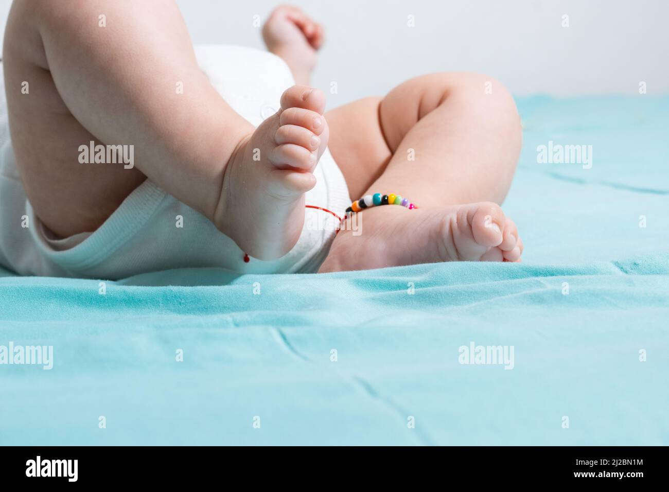pieds beaux et tendres d'un bébé de deux mois, allongé sur un lit avec des draps bleus, sur un de ses pieds elle a un bracelet de cheville et porte un whi Banque D'Images