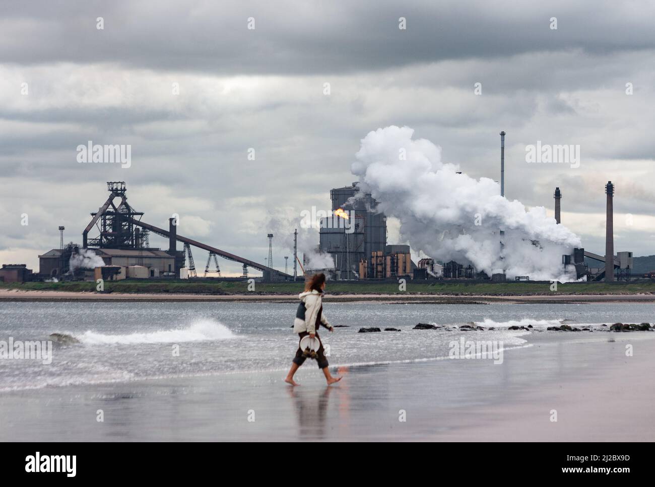 Femme marchant sur la plage de la gare du nord près de Seaton Carew avec des aciéries Redcar sur le côté sud de l'estuaire de Tess en arrière-plan. Angleterre. ROYAUME-UNI Banque D'Images