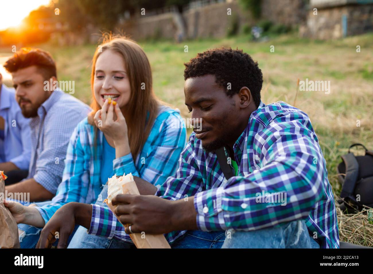 Les heureux étudiants multiethniques dans des vêtements décontractés sont assis sur l'herbe et de manger la restauration rapide Banque D'Images