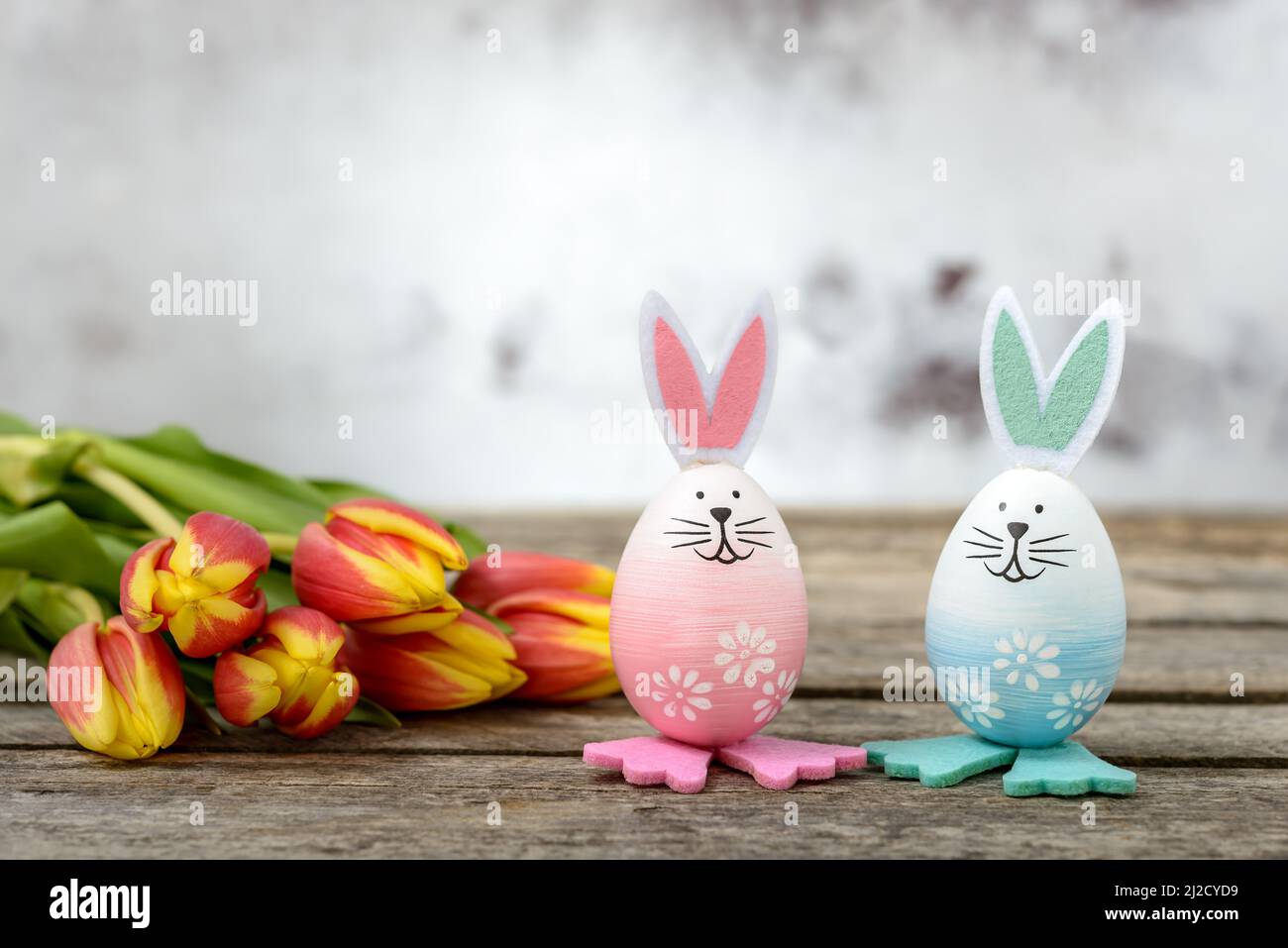 Deux œufs de Pâques avec un joli visage de lapin et des oreilles de lapin à côté de tulipes orange-jaune sur une table rustique en bois. Concept de Pâques. Banque D'Images