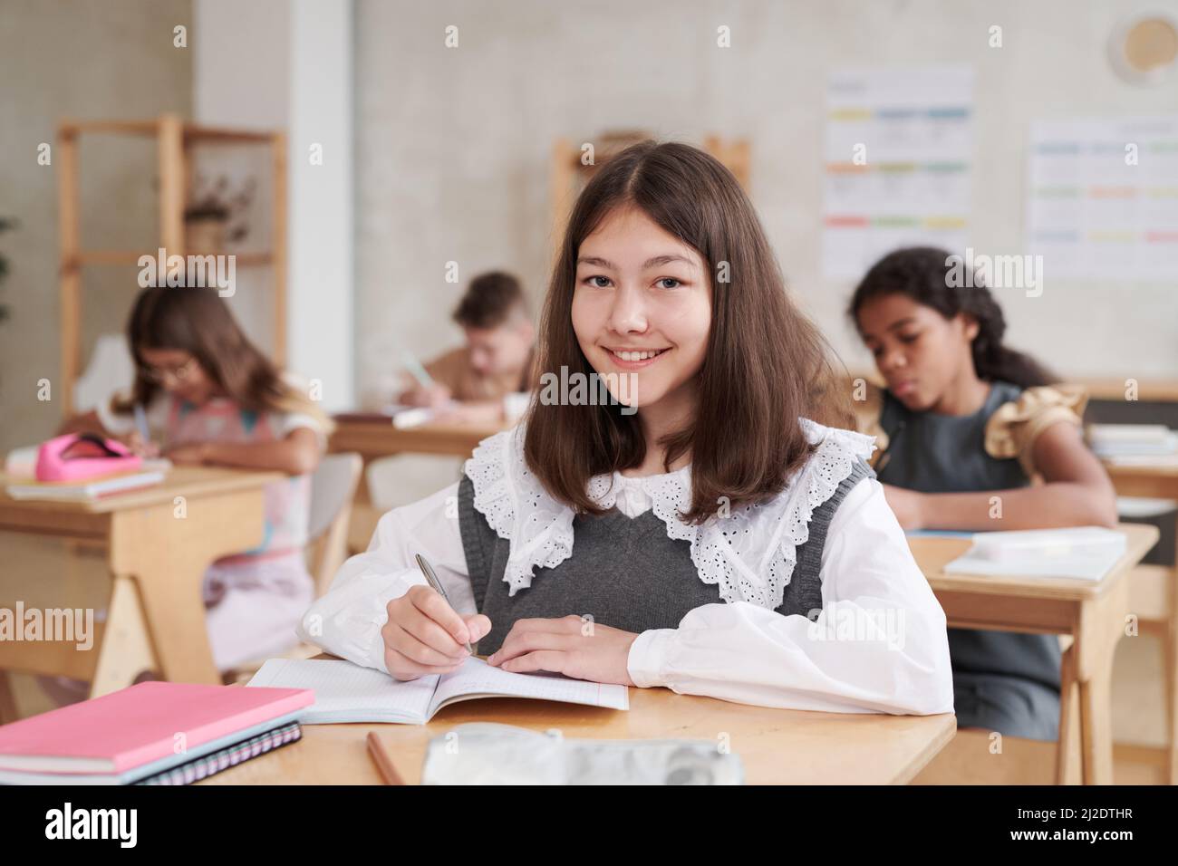 Portrait d'une écolière souriante portant un uniforme, assise au bureau dans une salle de classe, décor en bois, espace de photocopie Banque D'Images