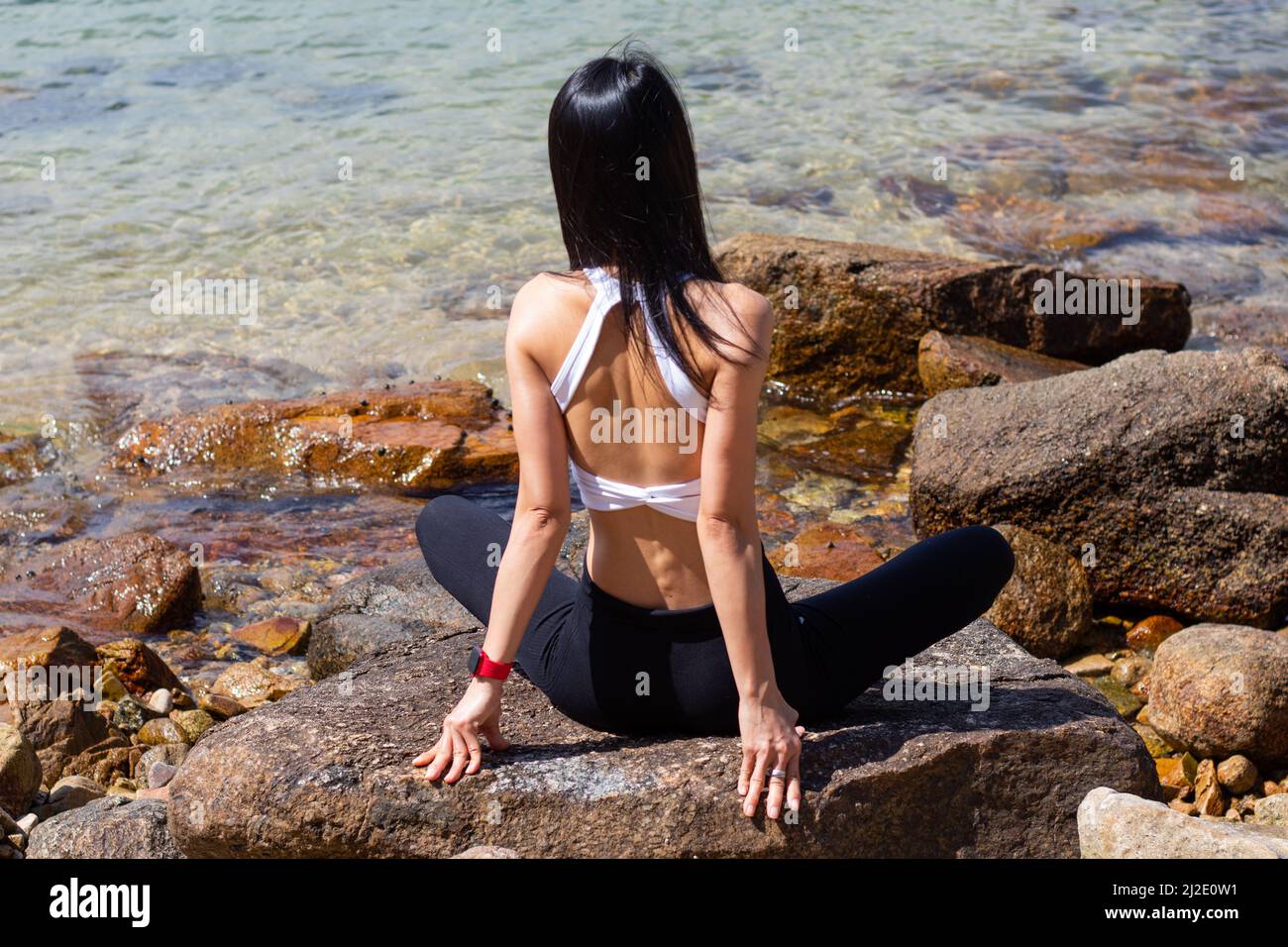 Une femme mince avec de longs cheveux noirs portant des vêtements de sport noirs et blancs est assise à pattes croisées sur des rochers surplombant la mer Banque D'Images