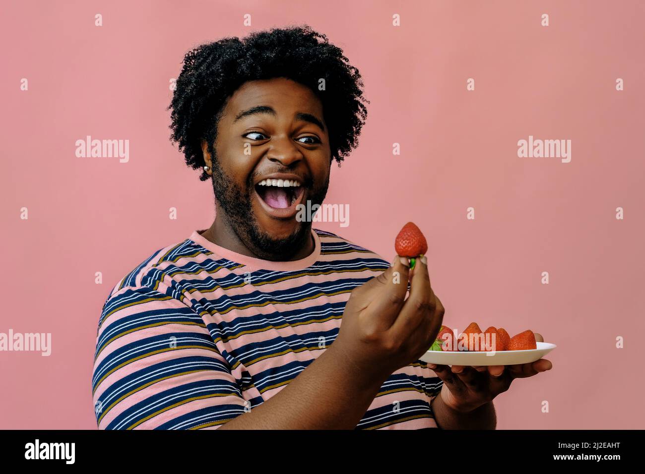 jeune homme afro-américain heureux mangeant des fraises dans le studio sur fond rose modèle masculin Banque D'Images