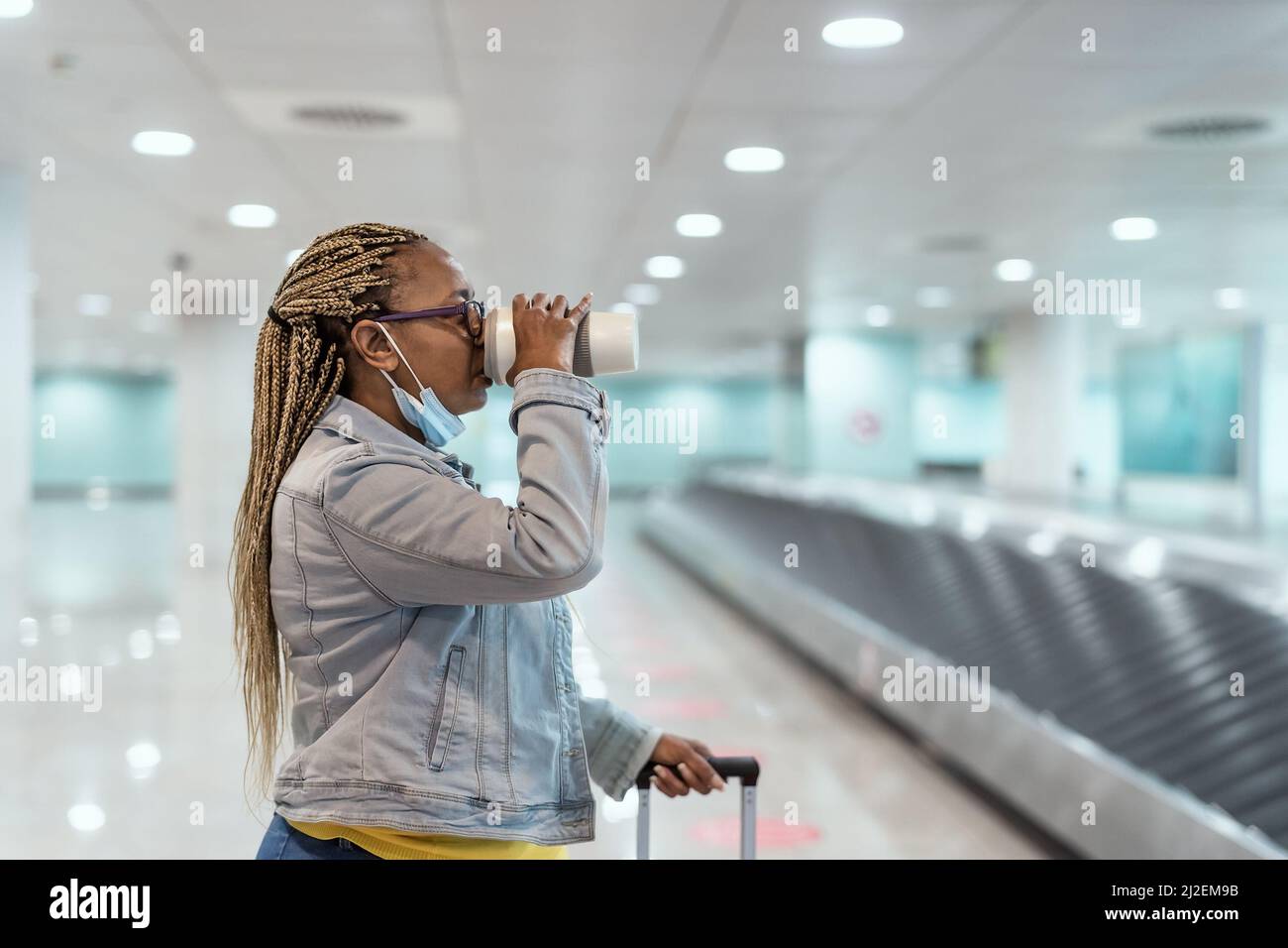 Femme africaine âgée marchant dans le terminal de l'aéroport avec ses bagages tout en buvant une tasse de café Banque D'Images