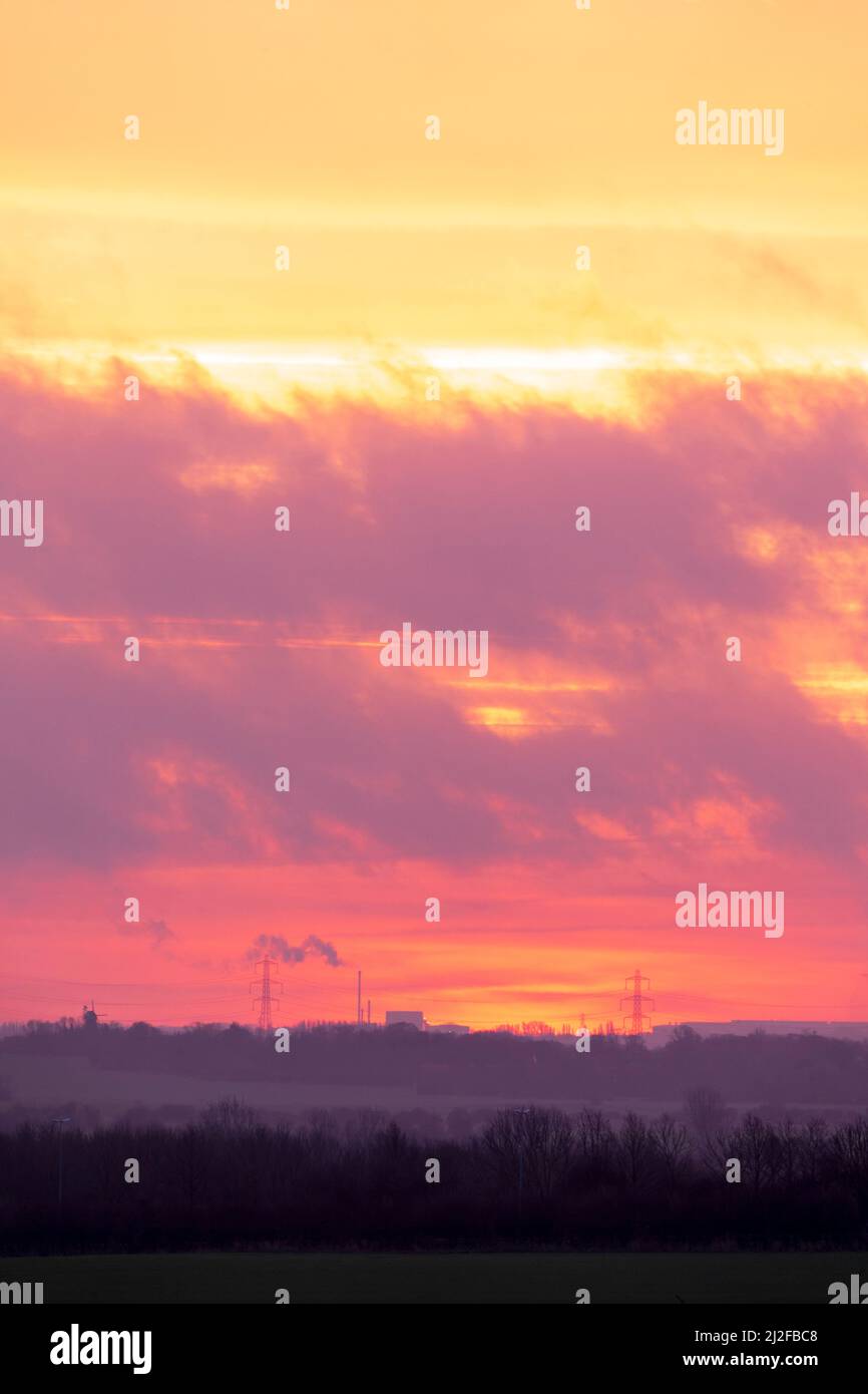 Ciel d'aube orange et jaune dominant un moulin à vent, pylônes électriques et une grande cheminée à fumer à l'horizon lointain. Arbres au premier plan. Banque D'Images