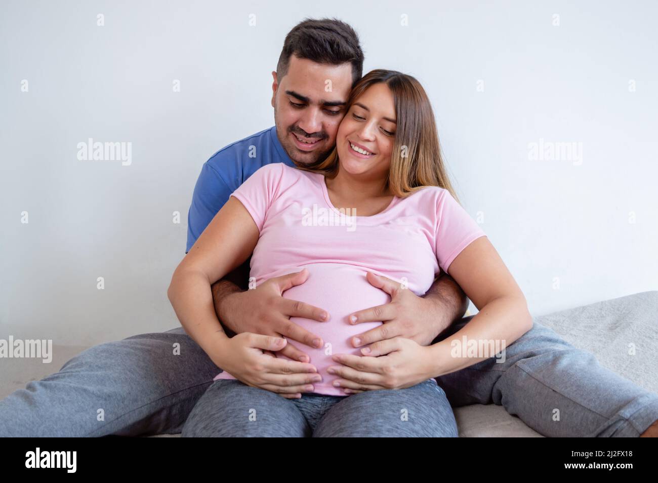 bonne femme enceinte caucasienne avec son mari assis sur le lit souriant et touchant bébé sur le ventre, faisant la thérapie de grossesse et la routine d'exercice. Banque D'Images