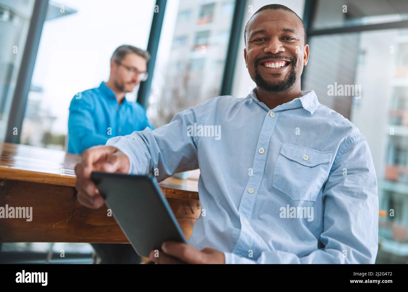 La technologie a vraiment simplifié notre façon de travailler. Portrait d'un homme d'affaires utilisant sa tablette avec un collègue en arrière-plan. Banque D'Images