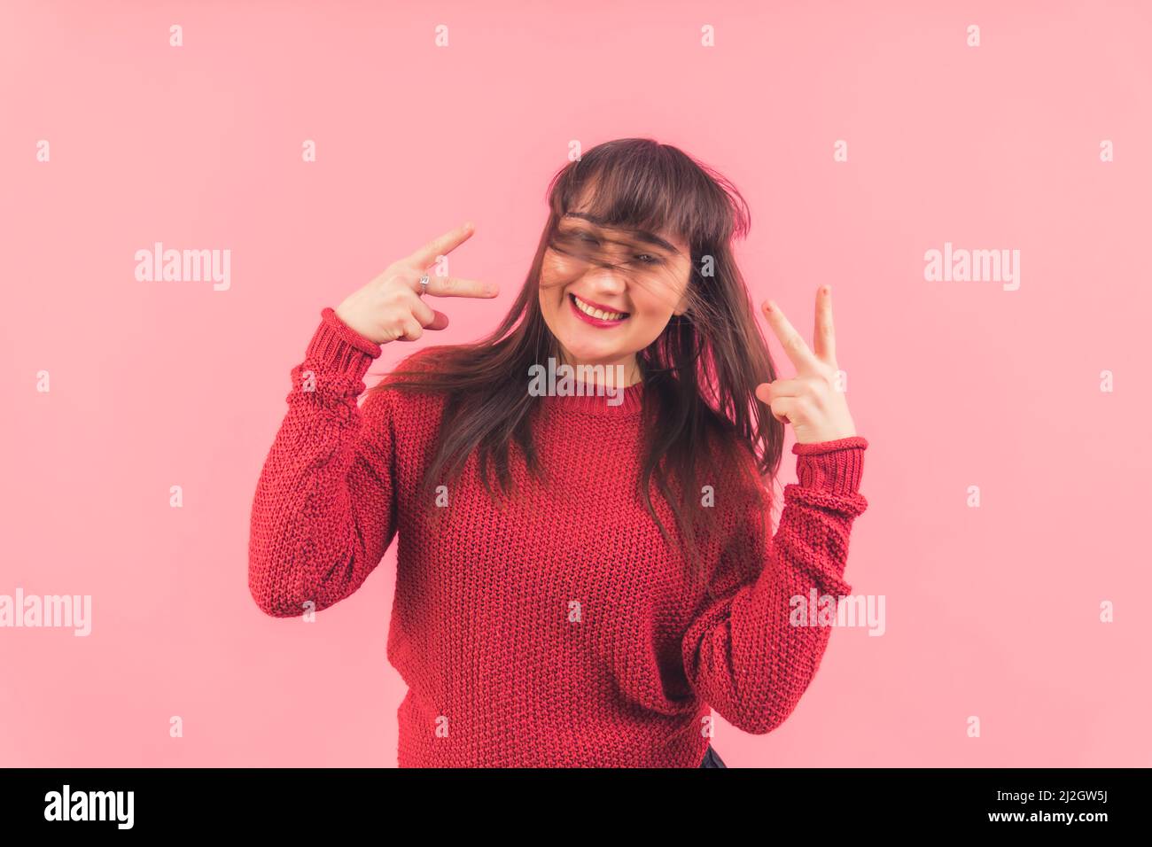 Joyeux brunette européenne drôle avec des cheveux couvrant son visage montrant le v-signe à la caméra et sourire. Fond rose, prise en studio. Photo de haute qualité Banque D'Images