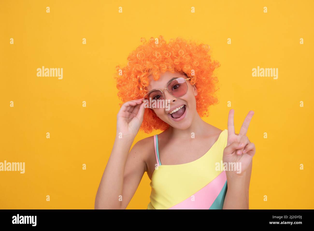 joyeux enfant dans des lunettes de soleil et un maillot de bain portant des cheveux de perruque orange montrant le geste de paix, la paix Banque D'Images