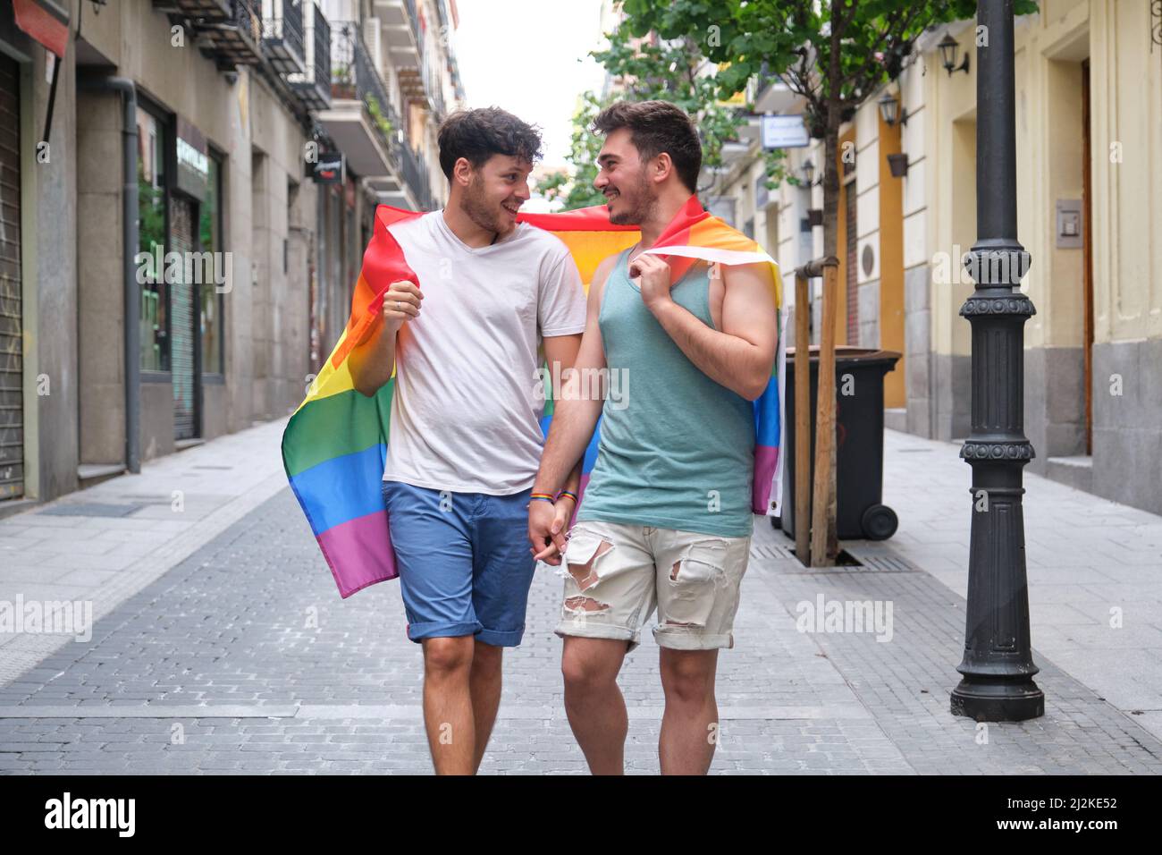 Couple gay marchant enveloppé dans un drapeau LGBT, regardant l'un l'autre et souriant. Banque D'Images