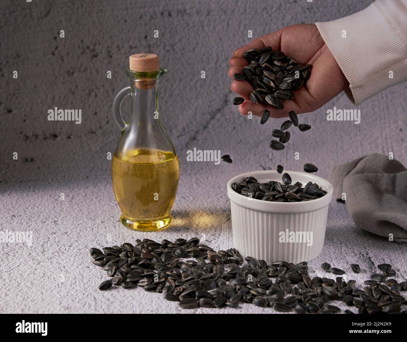 La main d'une femme anonyme verse des graines de tournesol dans un bol en céramique blanche et des graines de tournesol en vrac sur une table avec une bouteille en verre d'huile de tournesol Banque D'Images