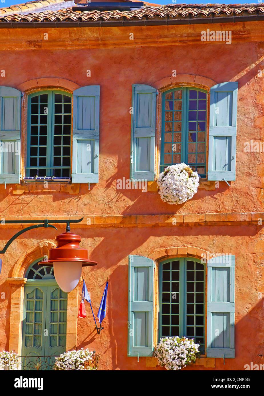 Vue sur la façade traditionnelle de la maison murale ocre en pierre naturelle, arrangements floraux, volets en bois de la vieille fenêtre - Hôtel de ville Roussillon, France Banque D'Images
