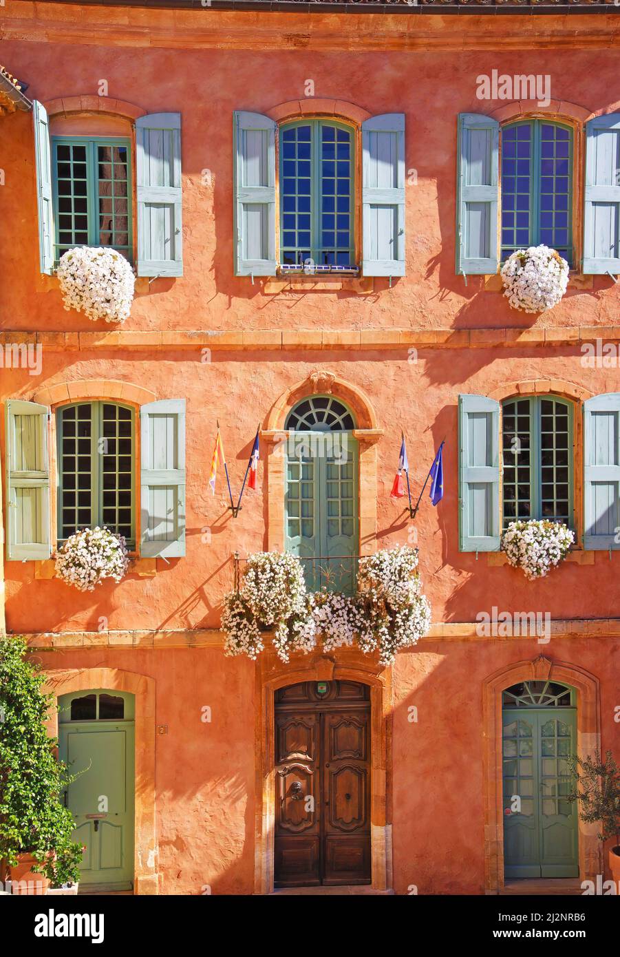 Vue sur la façade traditionnelle de la maison murale ocre en pierre naturelle, arrangements floraux, volets en bois de la vieille fenêtre - Hôtel de ville Roussillon, France Banque D'Images