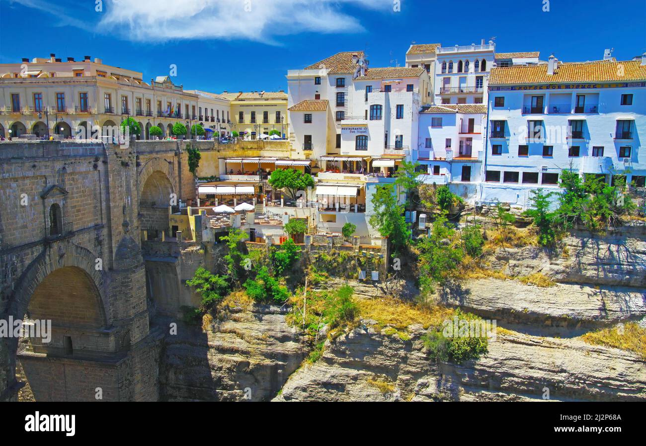 Vue au-delà du pont de pierre médiéval sur l'ancien village en haut de la falaise avec restaurant à flanc de falaise, ciel bleu - Ronda, Espagne Banque D'Images