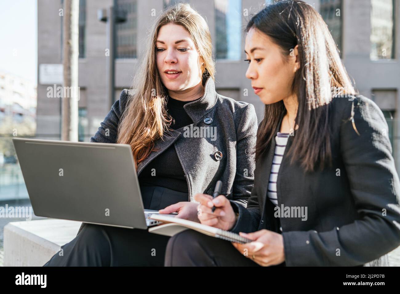 Femme en vêtements d'extérieur utilisant netbook tout en travaillant avec des collègues asiatiques écrivant dans le bloc-notes et expliquant les détails du projet Banque D'Images
