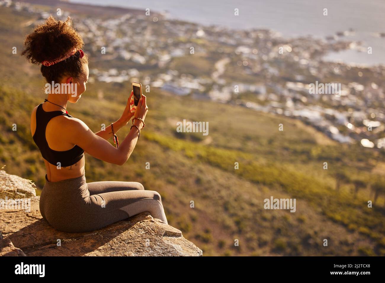 Voyons combien de goûts celui-ci va obtenir. Photo d'une femme prenant des photos avec son téléphone portable tout en étant assise sur une falaise de montagne. Banque D'Images