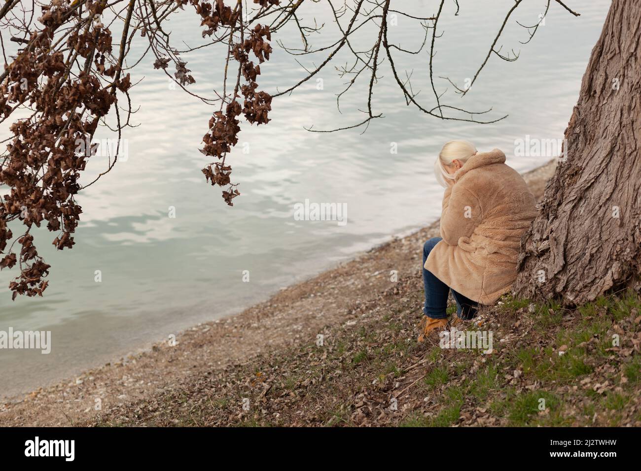 Femme blonde en manteau assise sous l'arbre au bord du lac, pleurant seule Banque D'Images