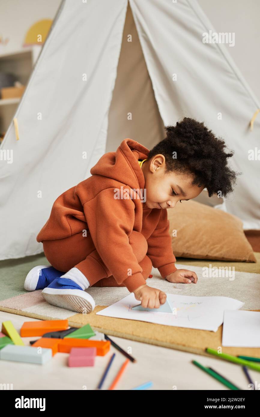 Portrait vertical de tout-petit garçon mignon dessin sur le sol dans la chambre des enfants tout en étant vêtu d'une tenue neutre tendance Banque D'Images