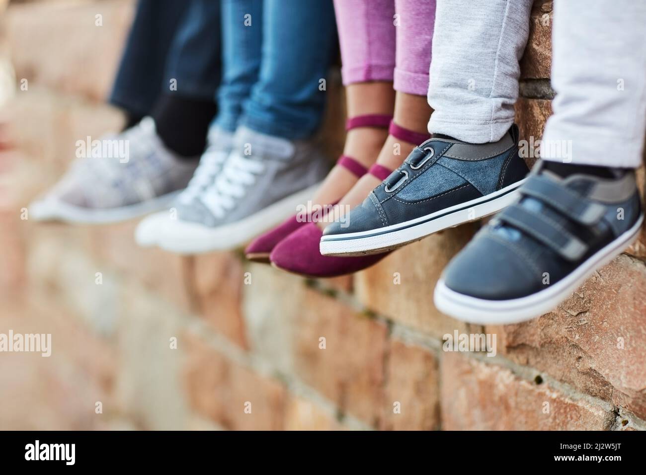 Des petits pieds qui vont dans de grands endroits. Photo courte d'enfants d'école élémentaire méconnus assis sur un mur de briques à l'extérieur. Banque D'Images