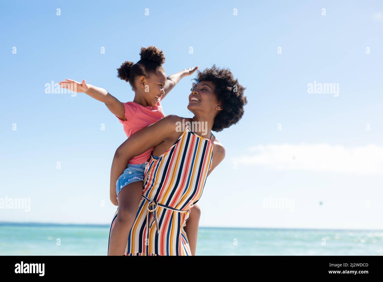 Bonne femme afro-américaine regardant sa fille avec les bras étendus sur son dos à la plage Banque D'Images