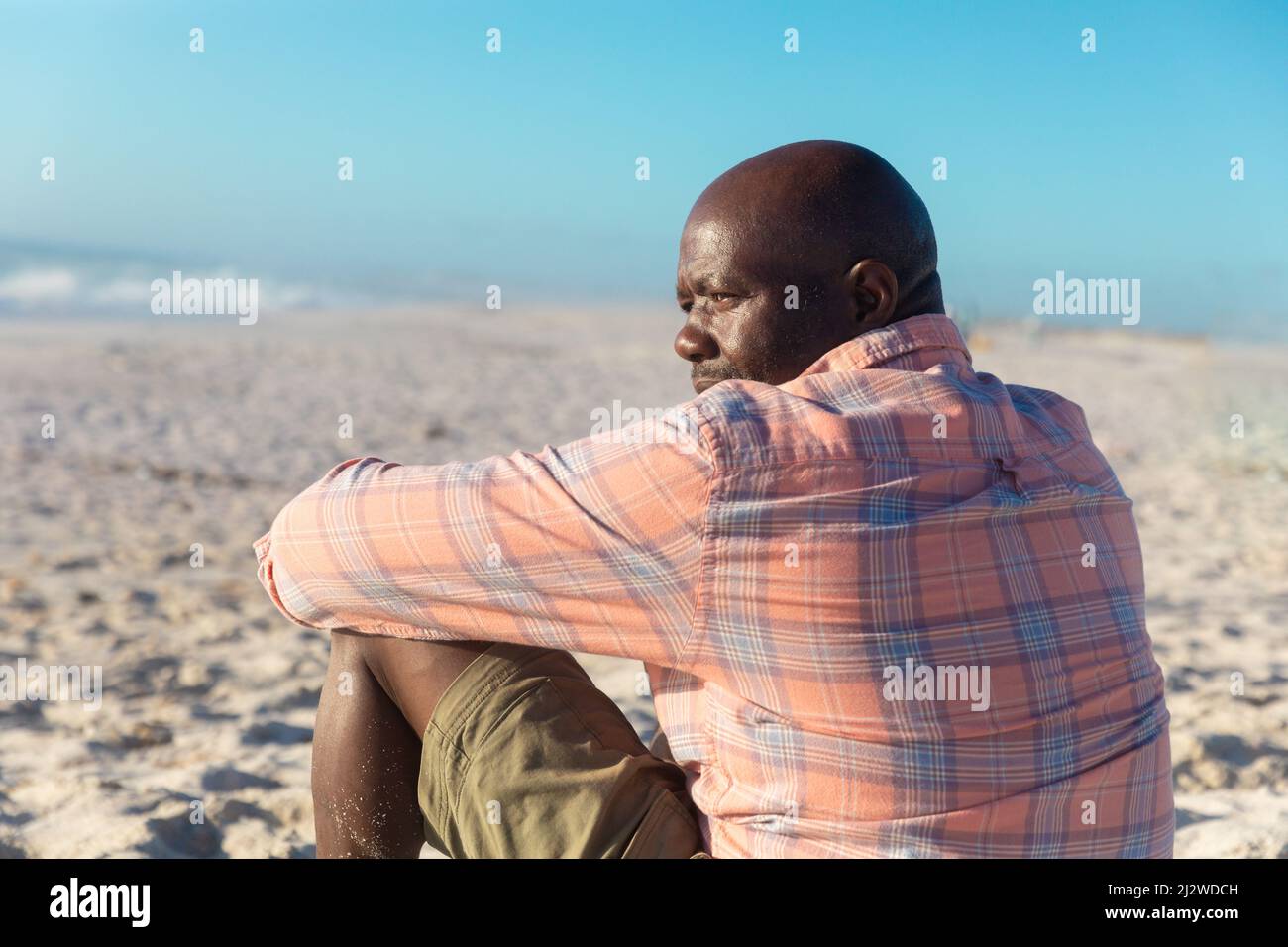 Vue arrière de l'homme senior afro-américain à la retraite qui regarde loin tout en étant assis à la plage par beau temps Banque D'Images