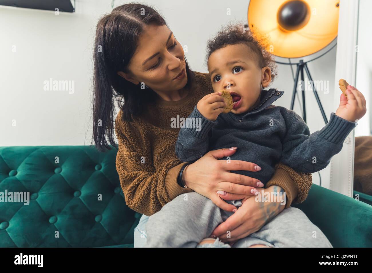 Afro-américain mignon petit garçon maudit dans les genoux de sa mère manger un cookie assis sur le canapé vert studio tourné moyen plein plan blanc arrière-plan copie espace. Photo de haute qualité Banque D'Images