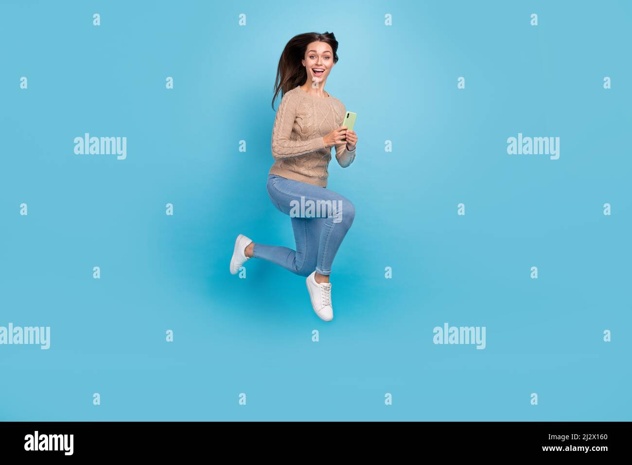 Photo de femme excitée saut tenir téléphone lire après courir vêtements beige chandail jeans baskets isolé couleur bleu fond Banque D'Images