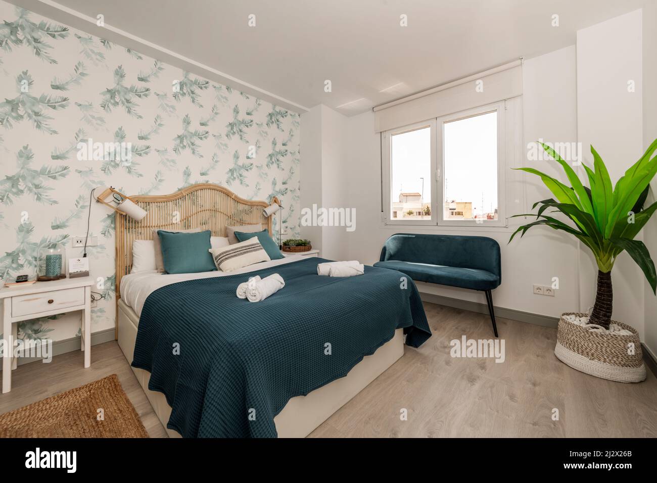 Chambre avec lit double et tête de lit en osier, murs avec papier peint décoratif à motifs végétaux, tapis en fibres naturelles, couverture bleue et natchi Banque D'Images