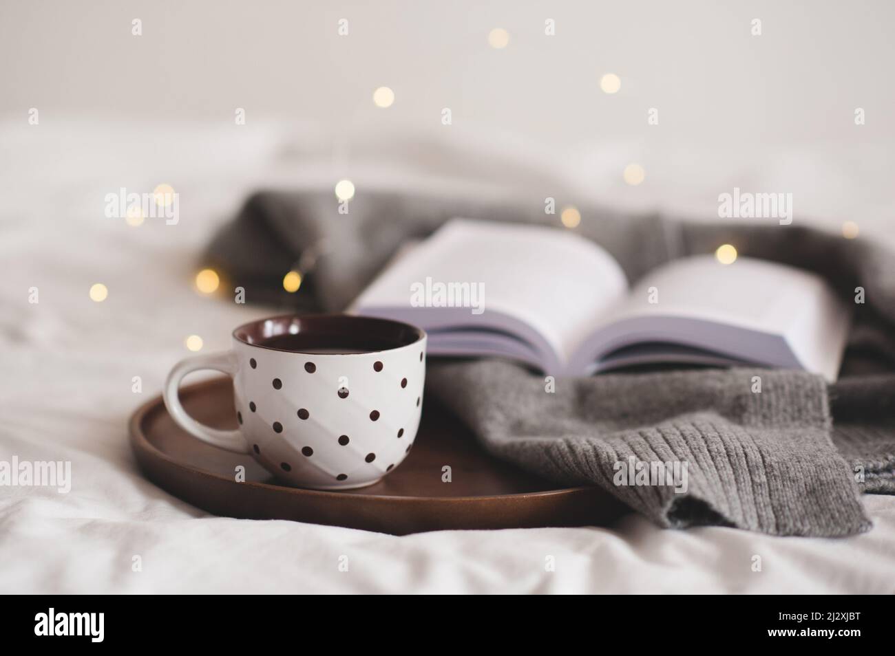 Tasse de café avec motifs à pois restant sur un plateau en bois avec textile tricoté et livre en papier ouvert sur des lumières phosphorescentes dans la chambre de près. Bonjour Banque D'Images