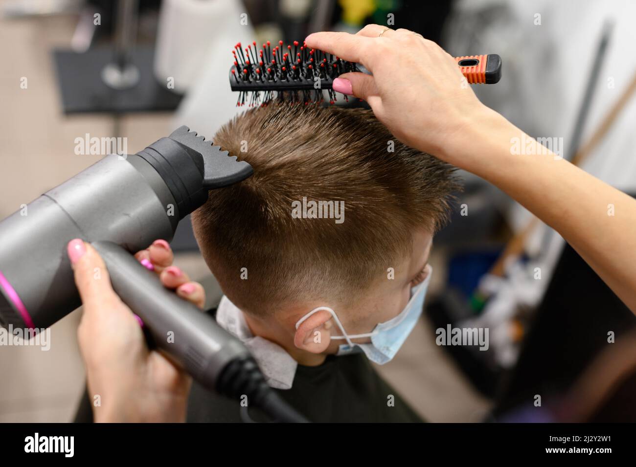 Le coiffeur sèche et coiffe les cheveux à l'aide d'un sèche-cheveux et d'un peigne pour un garçon d'école, visite au coiffeur pendant un coronavirus Banque D'Images