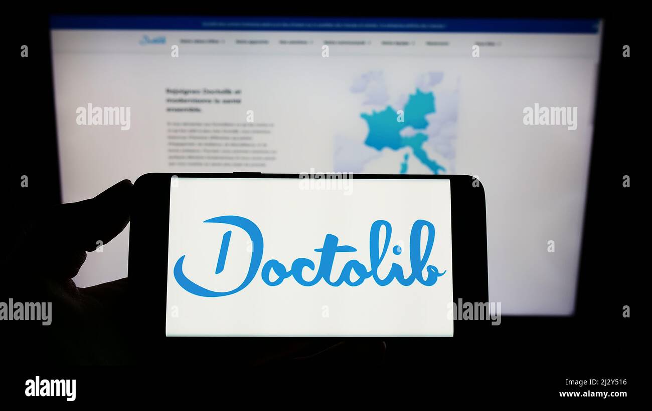 Personne tenant un téléphone portable avec le logo de la société française de soins de santé Doctolib SAS à l'écran en face de la page web d'affaires. Mise au point sur l'affichage du téléphone. Banque D'Images