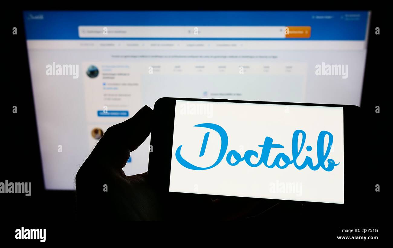 Personne tenant un téléphone portable avec le logo de la société française de santé Doctolib SAS à l'écran en face de la page web d'affaires. Mise au point sur l'affichage du téléphone. Banque D'Images