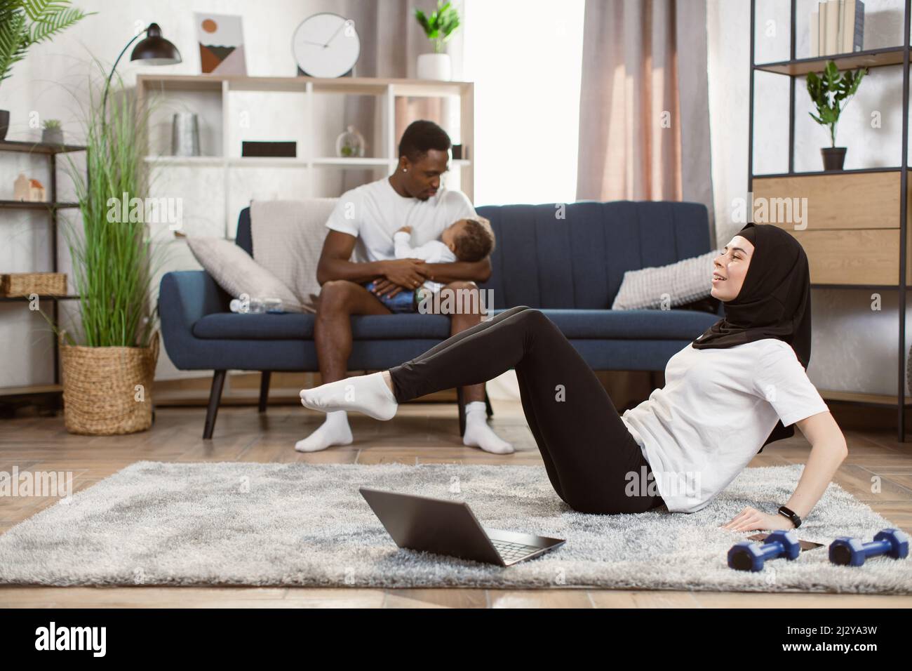 Une femme musulmane arabe dans un legging de sport et un t-shirt blanc  pratique le yoga dans le salon avec son mari et son fils afro-américains en  arrière-plan Photo Stock - Alamy