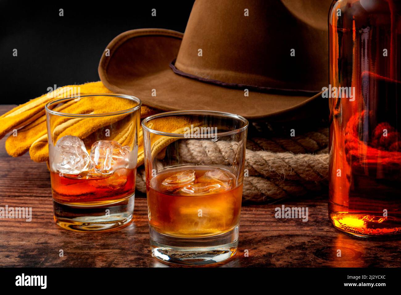 Le thème du concept de l'Ouest sauvage avec chapeau de cowboy, corde lasso, gants en cuir, deux verres de whisky sur les rochers et bouteille de bourbon sur table en bois Banque D'Images