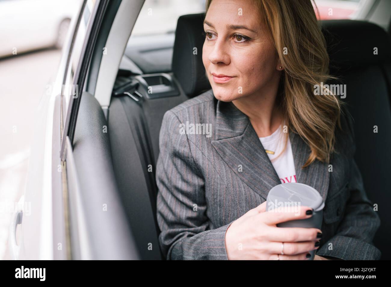 Une jeune femme d'affaires est assise dans le siège arrière d'une voiture, regardant par la fenêtre, buvant du café et se précipitant pour une réunion d'affaires. Banque D'Images