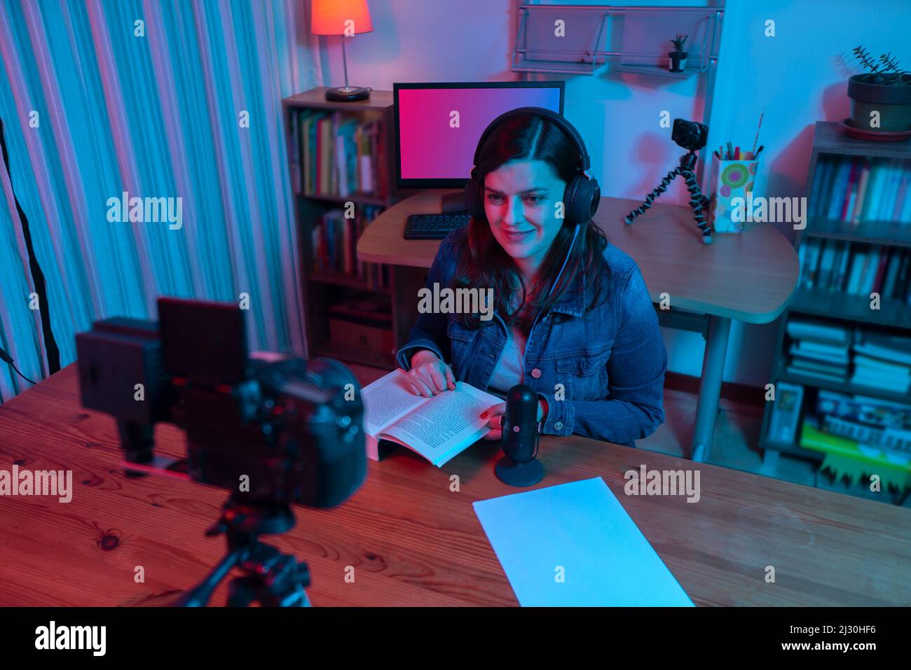 Belle femme hispanique devant une caméra vidéo enregistrant un blog dans son studio avec des lumières rouges et bleues à l'intérieur de sa maison. Diffusion de votre commen Banque D'Images