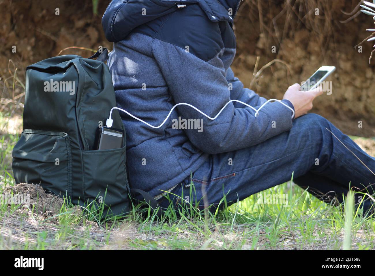 Un homme assis au sol, tient un smartphone dans ses mains et le charge avec une banque d'énergie noire sur fond de vue sur la nature. Appareil portable de voyage Banque D'Images