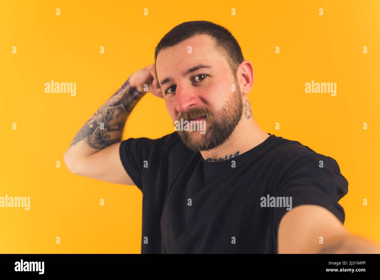 Homme européen attrayant avec une moustache à cheveux noirs courts barbe et des tatouages portant un T-shirt noir et prenant un selfie jaune fond studio prise de vue moyenne gros plan. Photo de haute qualité Banque D'Images
