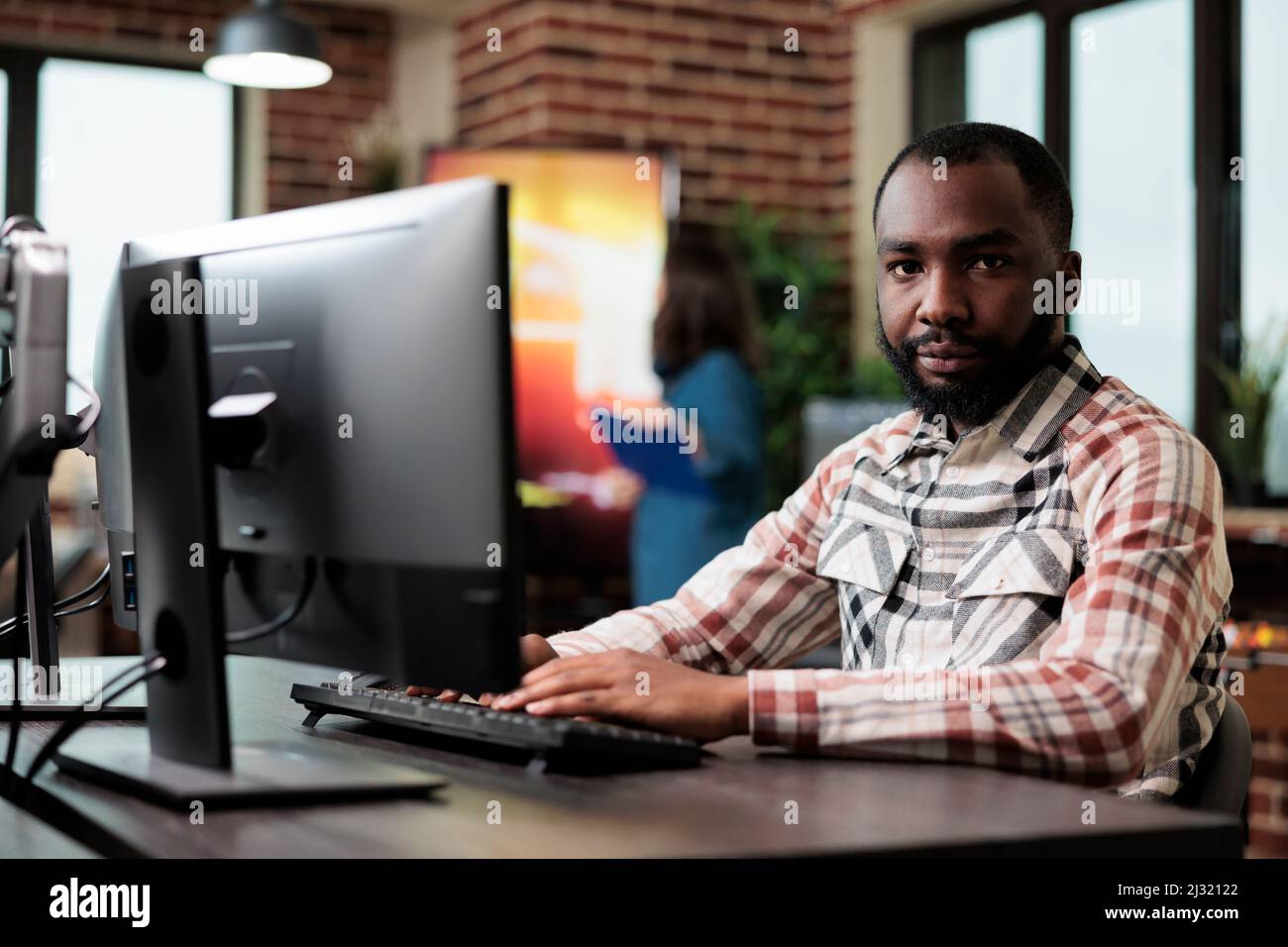Un employé d'une entreprise créative afro-américaine assis à un bureau dans un espace de travail tout en regardant la caméra. Agence adulte travailleur à l'ordinateur dans le studio de démarrage intérieur moderne. Banque D'Images
