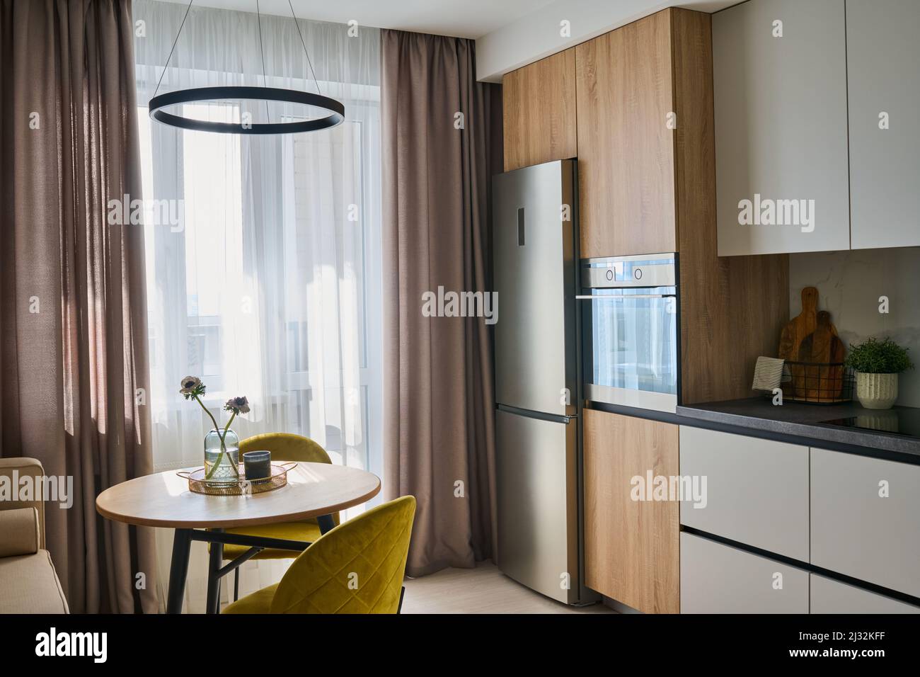 Table de cuisine ronde et deux chaises confortables de couleur jaune moutarde entre les fenêtres avec rideaux pastel et réfrigérateur Banque D'Images