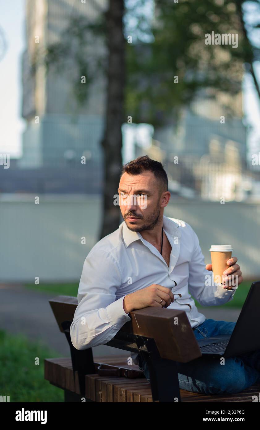 Homme attentionné dans le parc avec un ordinateur portable pour boire du café. Un jeune homme sur fond d'arbres verts, une chaude journée d'été ensoleillée. Lumière douce et chaude, gros plan. Banque D'Images