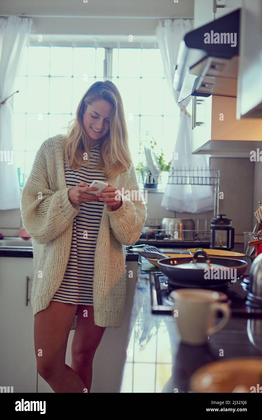 Le déjeuner est prêt, où êtes-vous. Photo courte d'une jeune femme attrayante qui envoie un SMS sur un téléphone portable tout en cuisinant dans sa cuisine. Banque D'Images