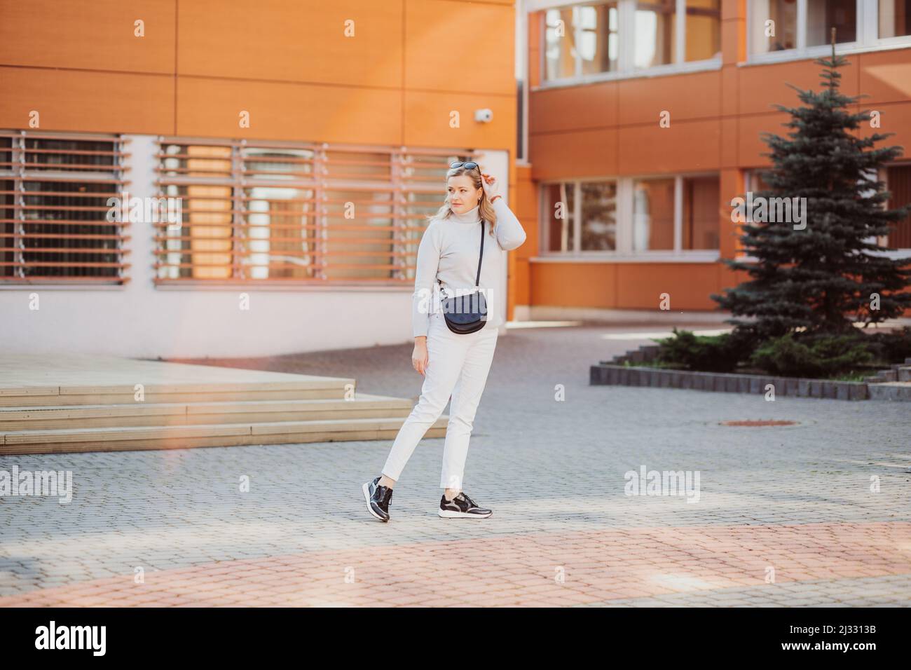 Blonde jolie femme élégante avec des lunettes de soleil, sac à bandoulière décontracté blanc vêtements marche route rue près du bâtiment épicéa. Banque D'Images