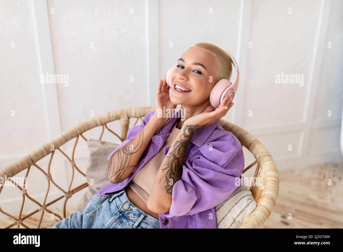 Jeune femme dans un casque sans fil se rafraîchissants dans un fauteuil en osier, écoutant de la musique et souriant à la caméra à la maison Banque D'Images