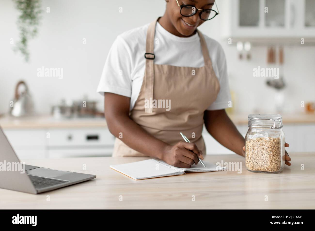 Une femme afro-américaine millénaire contente de lunettes et de tablier écrit une nouvelle recette avec un ordinateur portable dans la cuisine scandinave moderne Banque D'Images