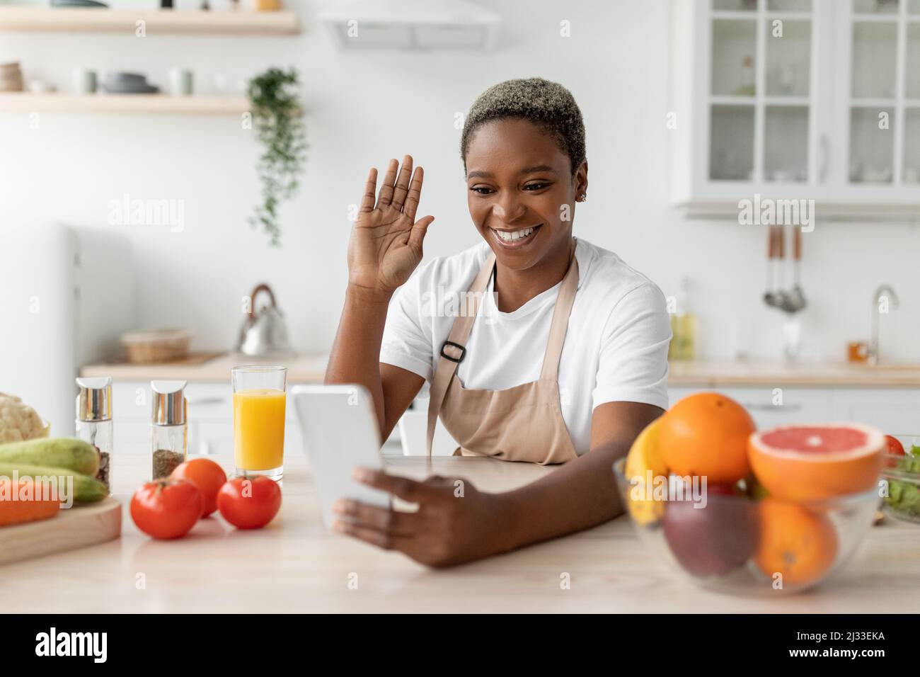 Femme noire millénaire souriante dans un tablier en agitant la main au téléphone, assis à table avec des légumes et des fruits dans la cuisine Banque D'Images