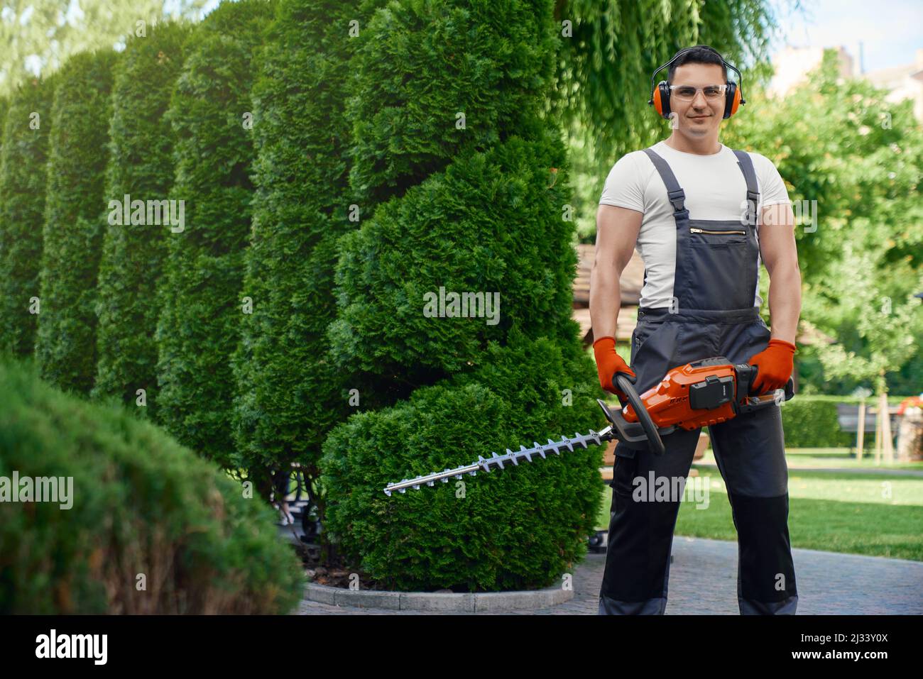 Jardinier caucasien compétent souriant et regardant la caméra tout en se tenant debout sur le jardin arrière et tenant le coupe-herbe électrique main. Homme utilisant un outil de jardinage pour couper la haie. Banque D'Images