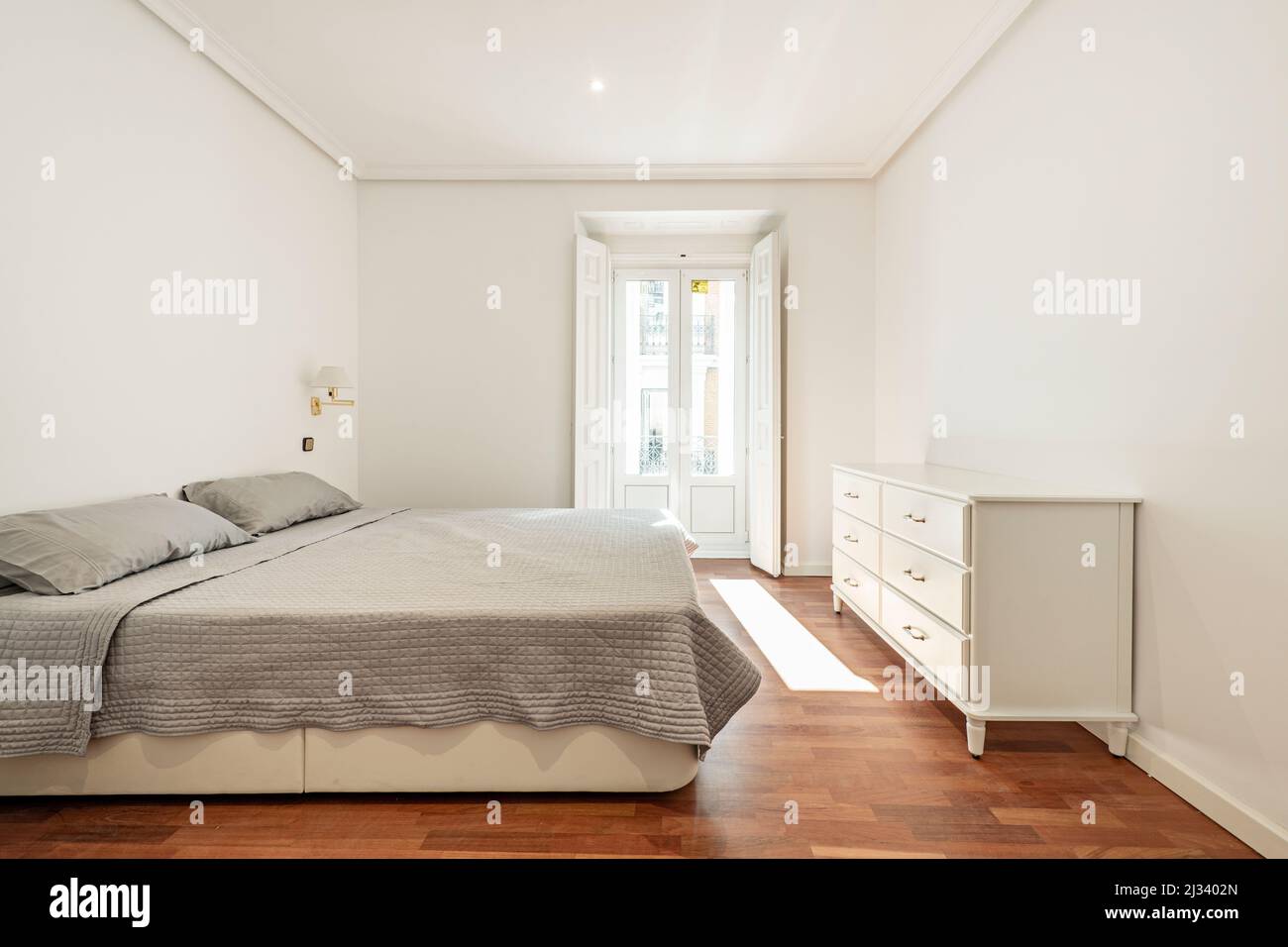 Chambre avec lit double et couvre-lit gris, commode en bois blanc et balcon avec volets en bois Banque D'Images