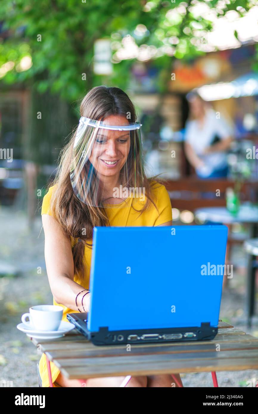 femme assise dans un café vide portant une visière de protection et travaillant sur un ordinateur portable Banque D'Images