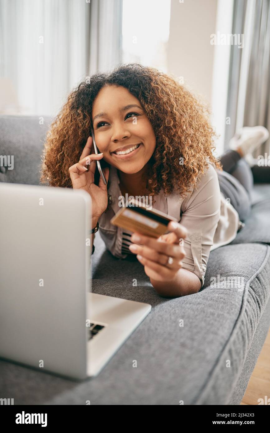 Shopping dans le confort de la maison. Photo d'une jeune femme joyeuse faisant des achats en ligne sur son ordinateur portable tout en parlant sur son téléphone portable à la maison. Banque D'Images